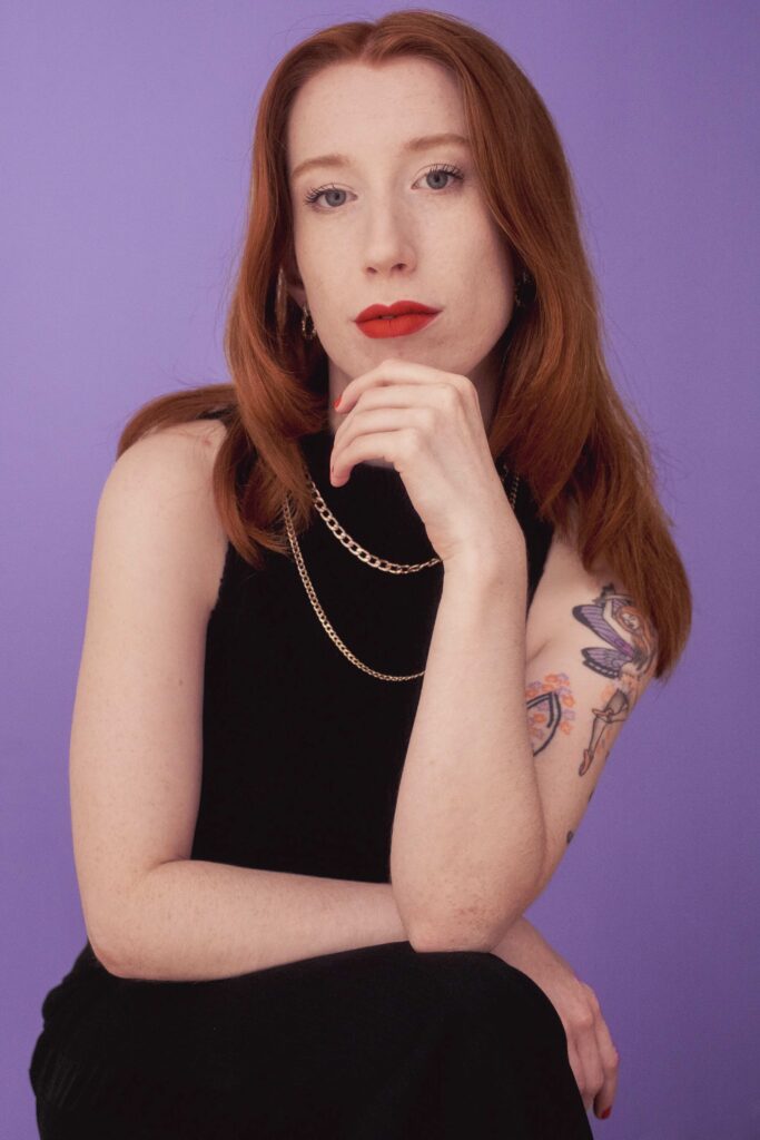 a woman with red hair and tattoos wearing black with gold chains looking at the camera
