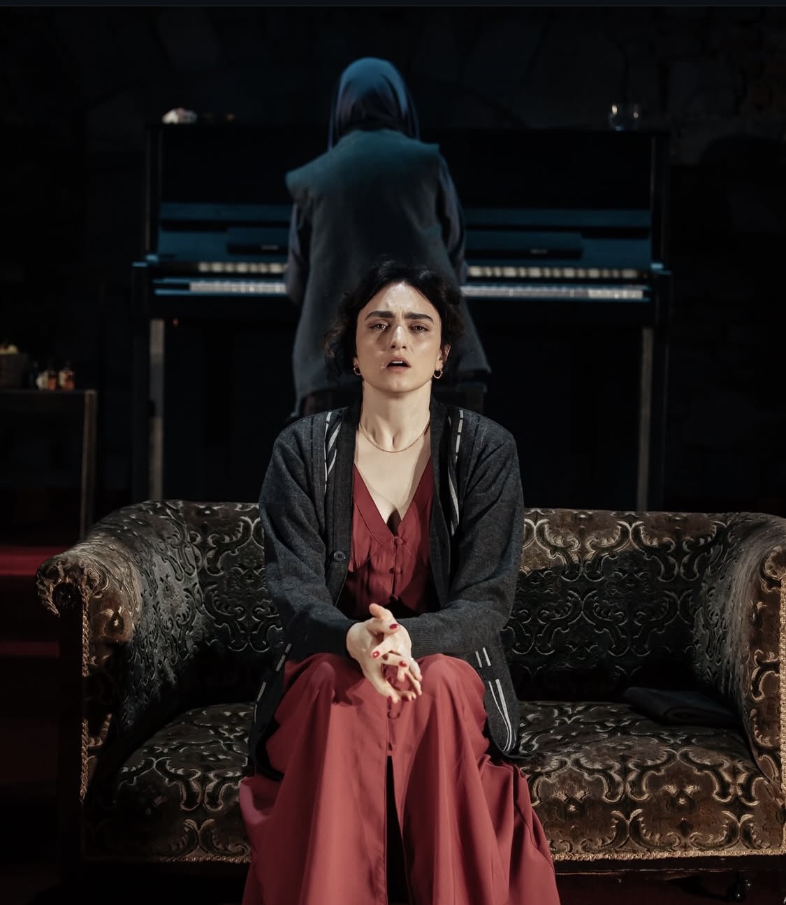 A white woman with black hair in a red dress sitting on a couch. A hooded figure sits at the piano in the background