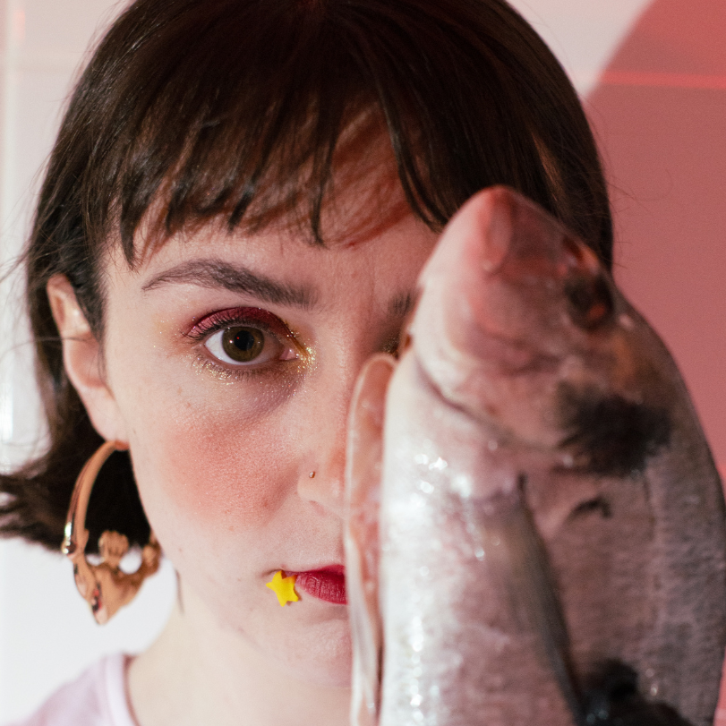 A white girl with short brown hair and a fringe and large cladagh earrings holding a fish up that covers half her face