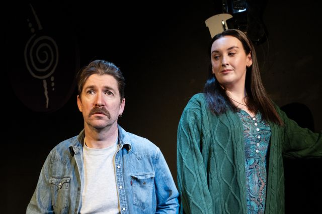 A white man and woman, both with dark hair standing beside each other. She wears a green cardigan and he wears a denim jacket with a white top
